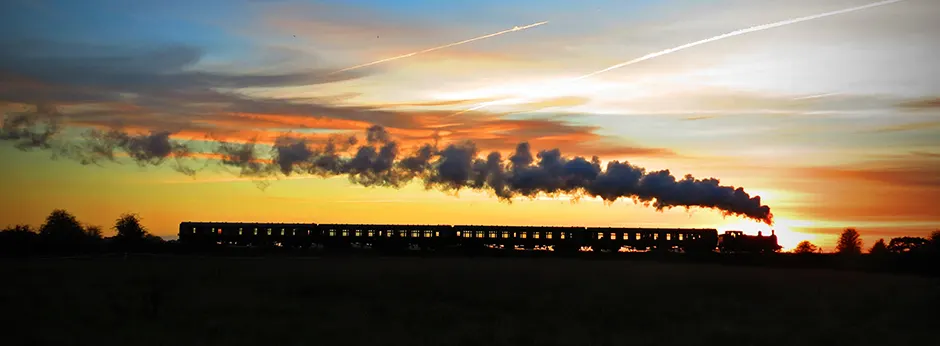 Steam train with illuminated carriages travelling through countryside at sunset with dramatic orange and golden sky.