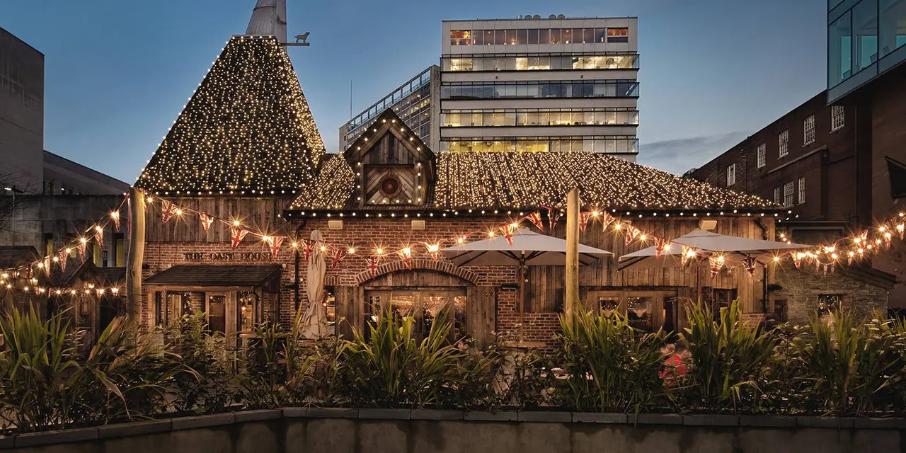 Tudor-style buildings with fairy lights and festive decorations beside a waterside courtyard at dusk.