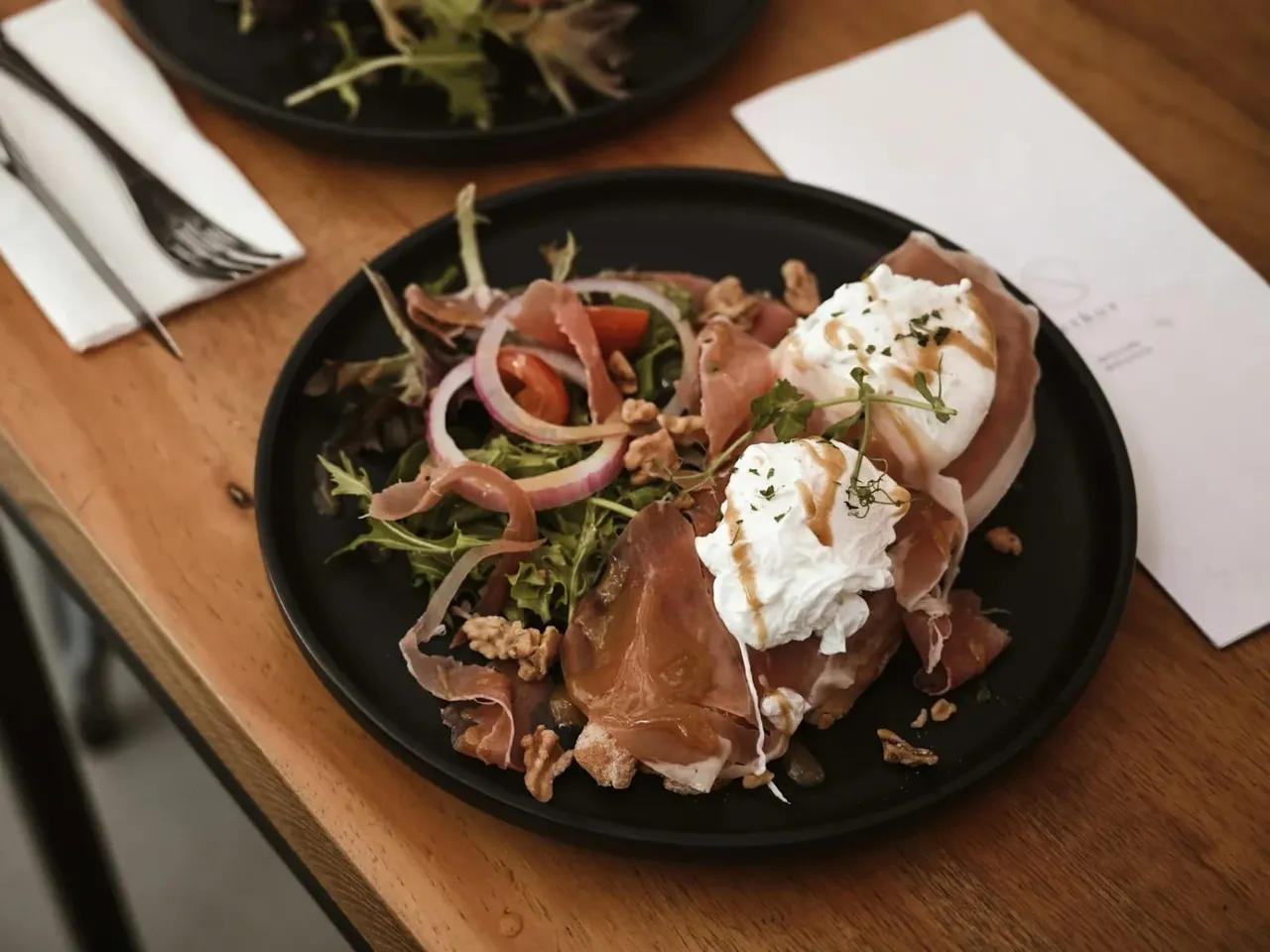 Plated dish with cured meat, burrata cheese, greens and pickled onions on a wooden table.