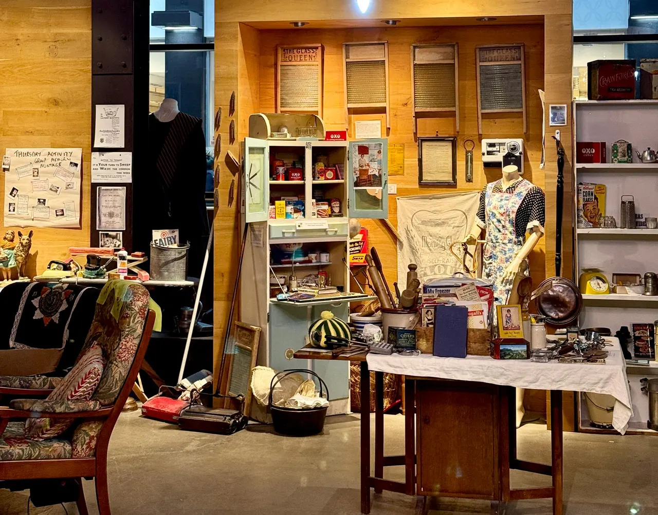 Museum display of domestic life and vintage items including furniture, kitchenware, clothing and memorabilia arranged in a recreated period room setting.