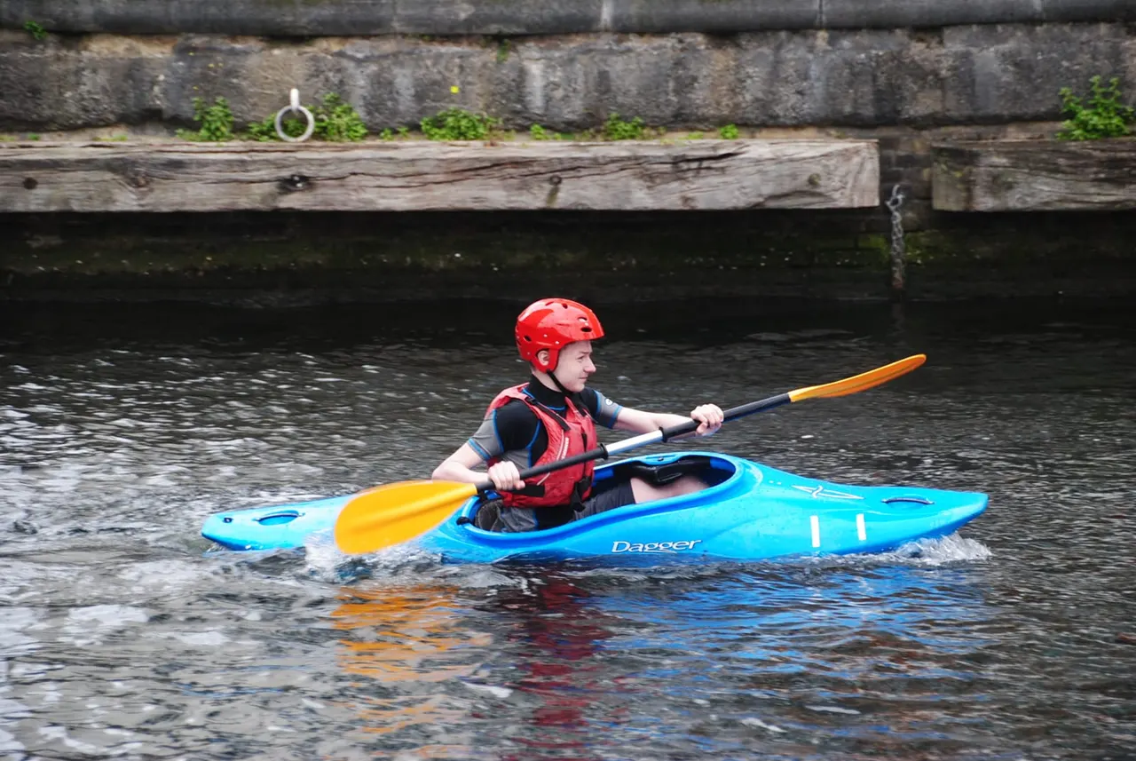 Child kayaking on a river in a blue kayak, wearing a red helmet and life jacket.