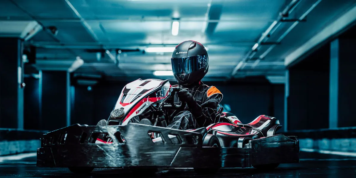 Person in helmet and racing suit driving go-kart indoors under fluorescent lights.