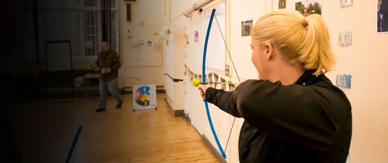 Woman aiming a bow and arrow at a target in an indoor activity space, with another person visible in the background.