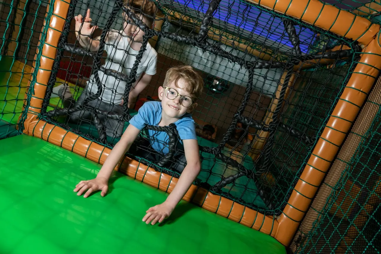 Child wearing glasses crawling through a large soft play structure with netting and padded sections.