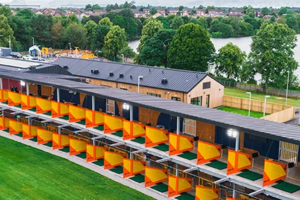 A row of bright yellow golf bays with covered shelters overlooks a river and tree-lined suburban landscape.