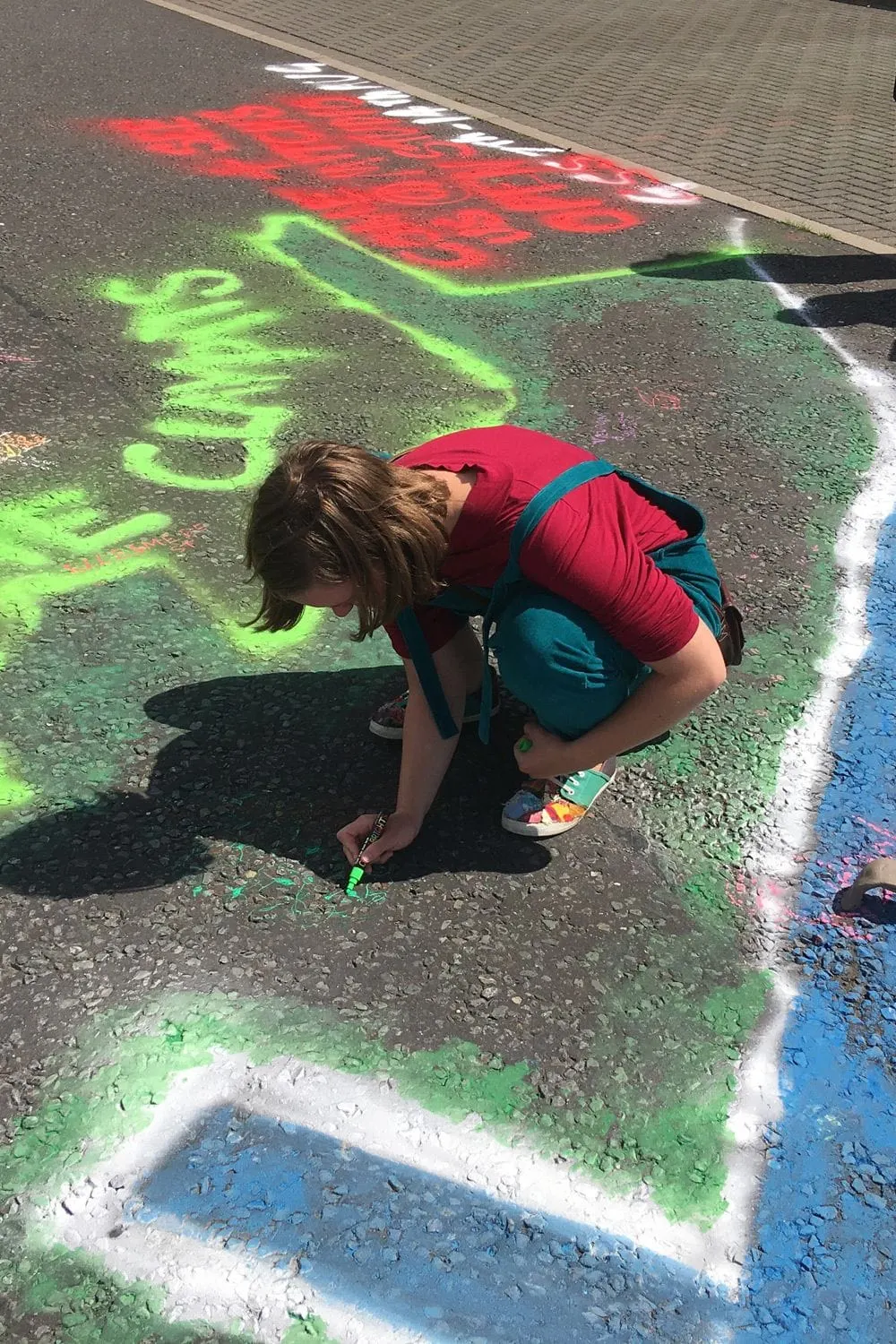 Child drawing with chalk on pavement, creating bright green and red artwork with white outlines.