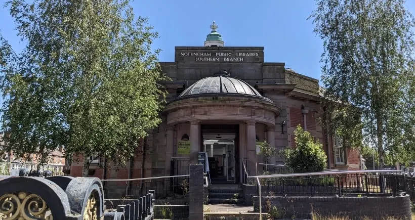 Historic red-brick library building with domed roof and classical architecture, surrounded by birch trees on a sunny day.