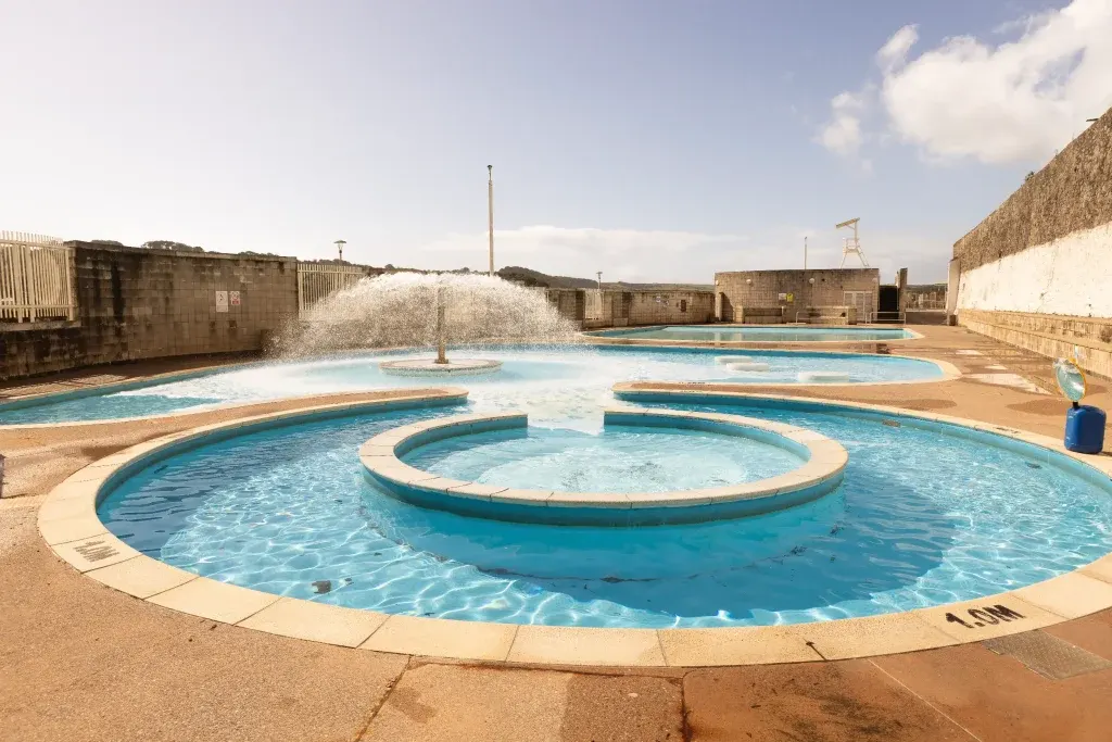Open-air thermal pools with turquoise water and central fountain feature, surrounded by stone buildings and pale sky.