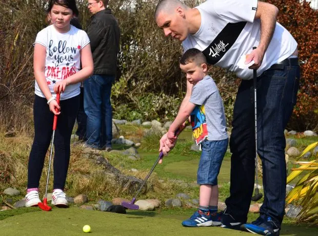 An adult instructing a young child on how to putt on a miniature golf course, with other visitors watching nearby.