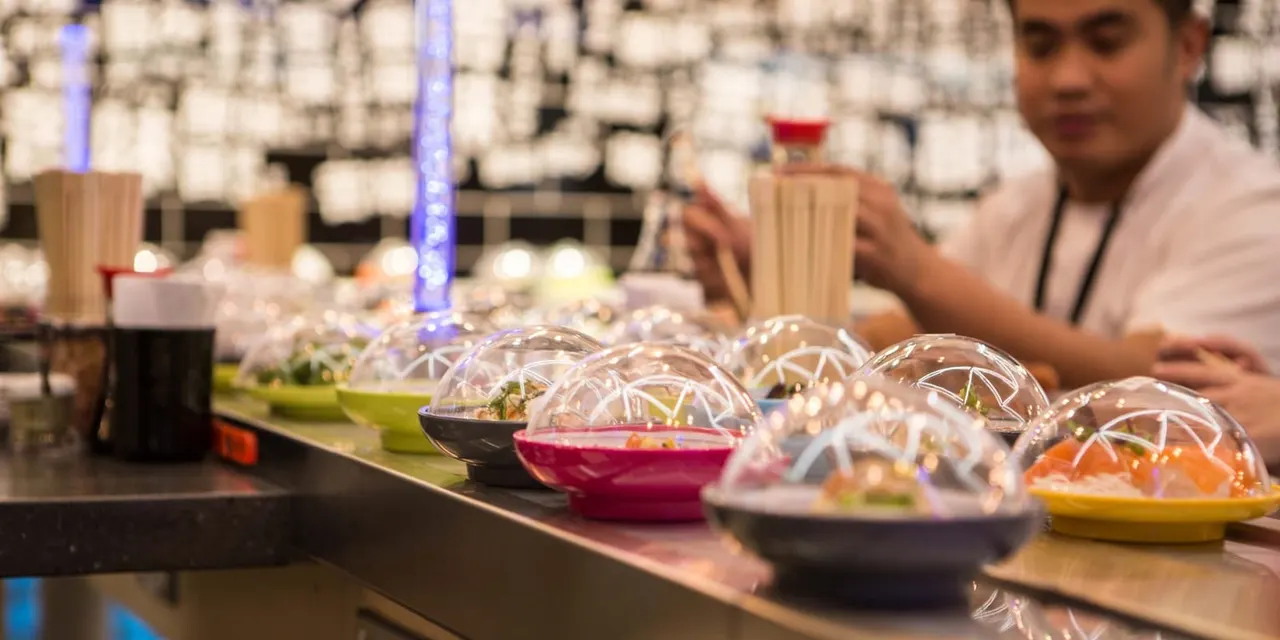 Plated dishes with glass domes line a counter at a fine dining restaurant, with a chef working in the background.