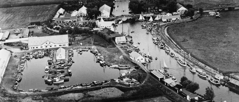 Aerial black and white photograph of a riverside leisure venue with moored boats, waterside buildings, and surrounding fields.
