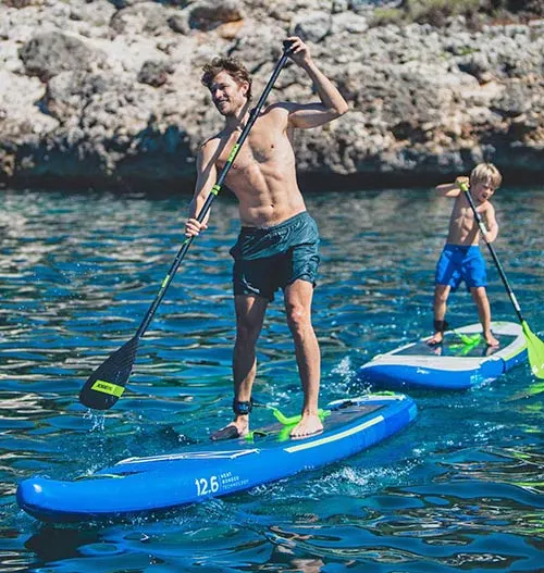 Man and child stand-up paddleboarding on blue boards in clear turquoise water near rocky cliffs.
