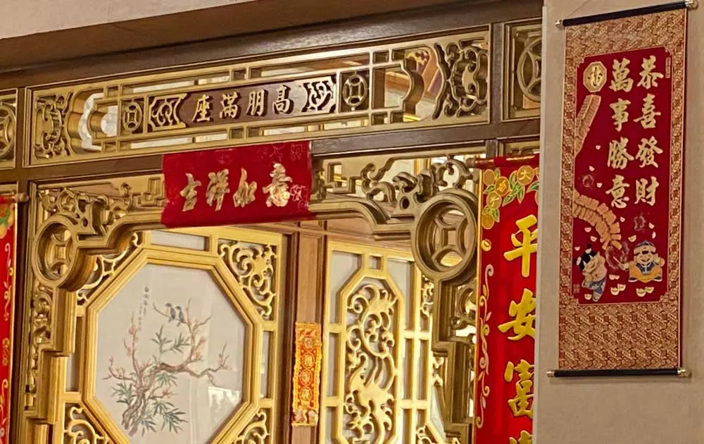 Ornate golden wooden panels with traditional Chinese carvings and red fabric banners with gold lettering decorating an interior wall.
