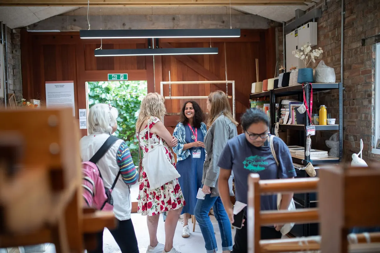 Visitors browse an indoor craft or retail space with exposed brick walls, shelving displaying textiles and homewares, and a staff member assisting customers.