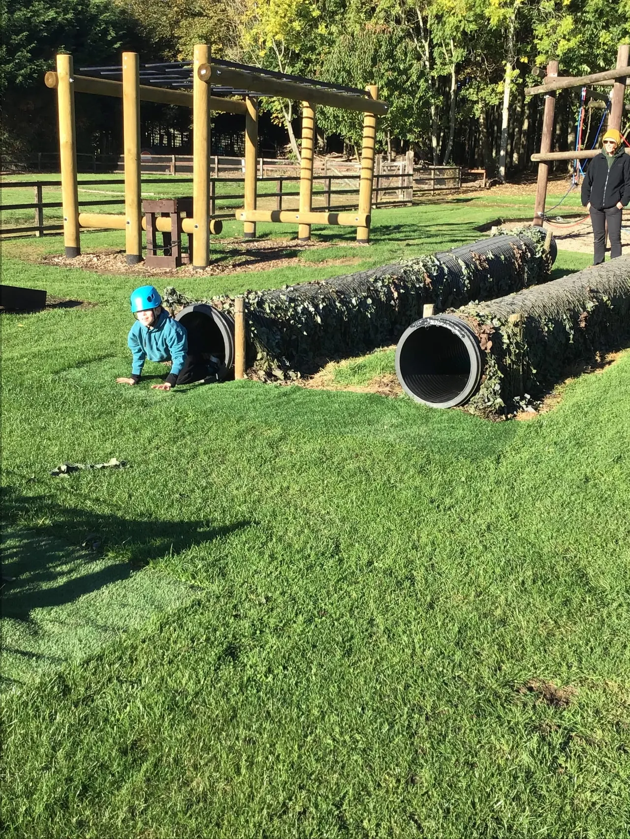 Child in blue helmet crawling through large pipe tunnel in outdoor adventure play area with wooden climbing structures and fencing.
