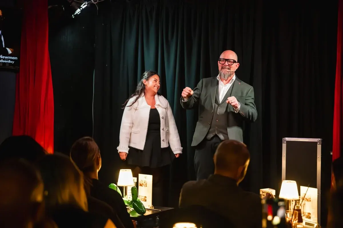 Two performers on stage with red and dark curtains behind them, engaging with an audience seated at tables in front.
