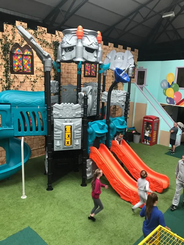 Children playing on a robot-themed indoor play structure with red slides in an indoor play centre.