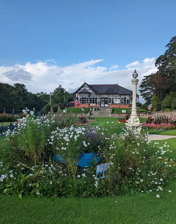 Half-timbered leisure pavilion with manicured gardens featuring white flowers, a memorial stone, and green lawns under blue sky.