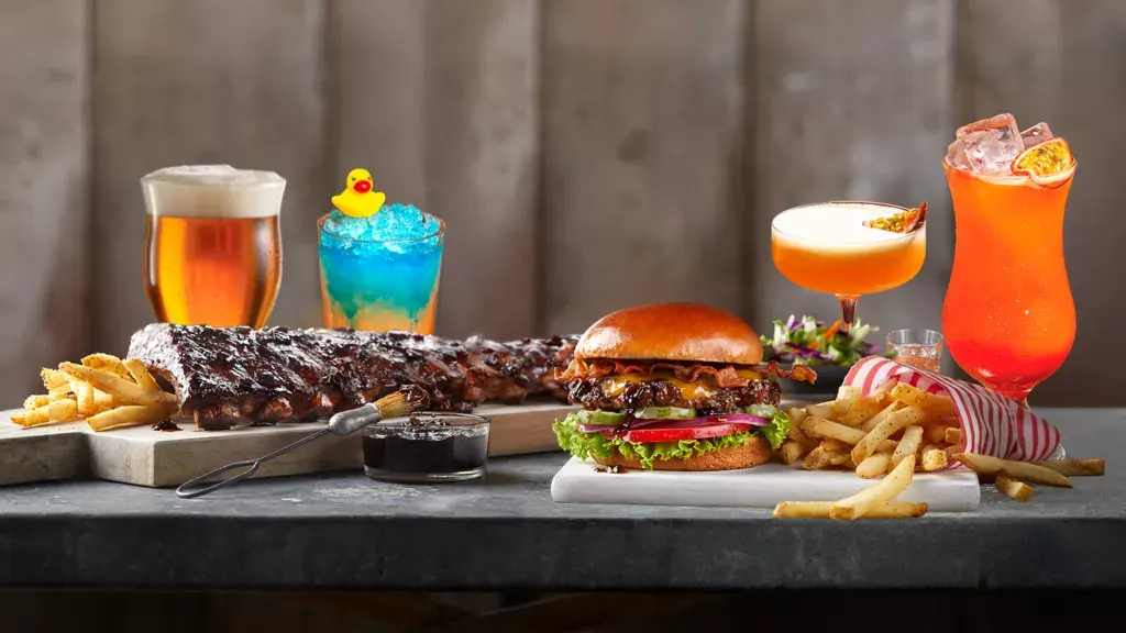 A spread of burgers, grilled meat, chips and colourful cocktails arranged on a dark counter against a wooden backdrop.