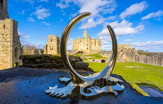 Modern curved sculpture framing a medieval castle and its grounds under blue sky.