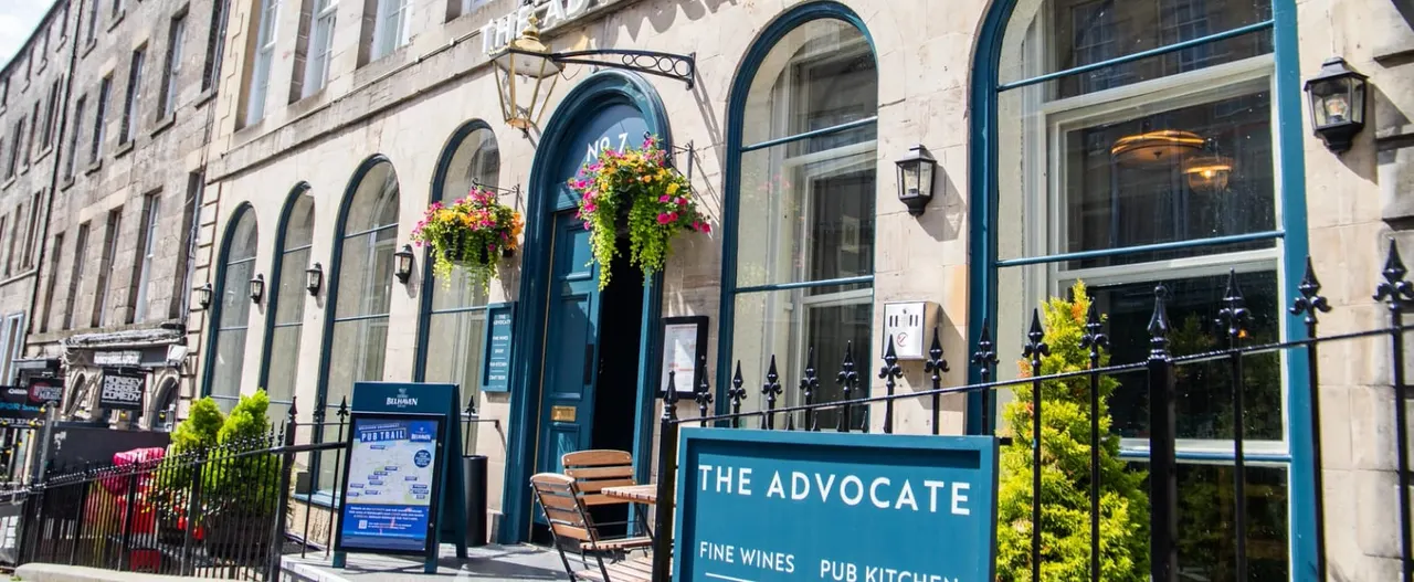 Historic stone building with blue-trimmed arched windows and doors, hanging flower baskets, and outdoor seating.
