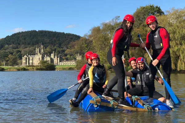 Group of people in red helmets and wetsuits paddling a blue raft on a river with a castle visible across the water.