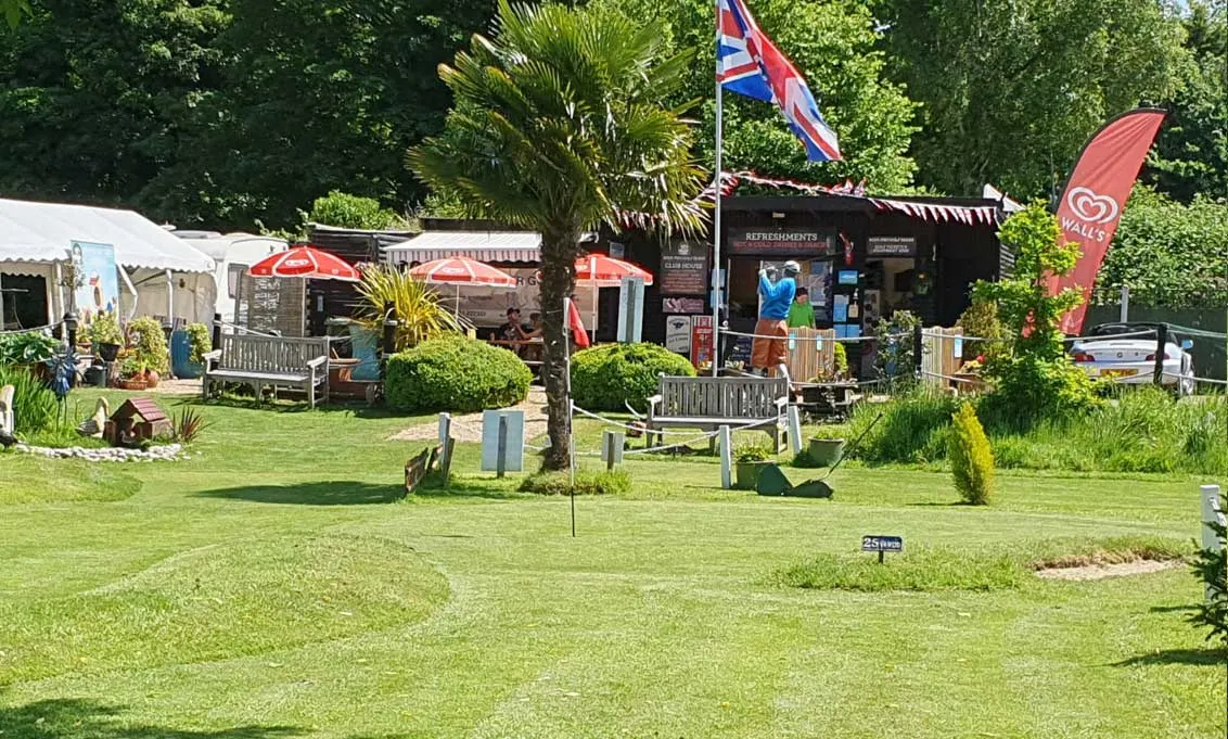 Mini golf course with putting green in foreground, refreshment hut and seating areas beneath umbrellas in background, surrounded by trees.