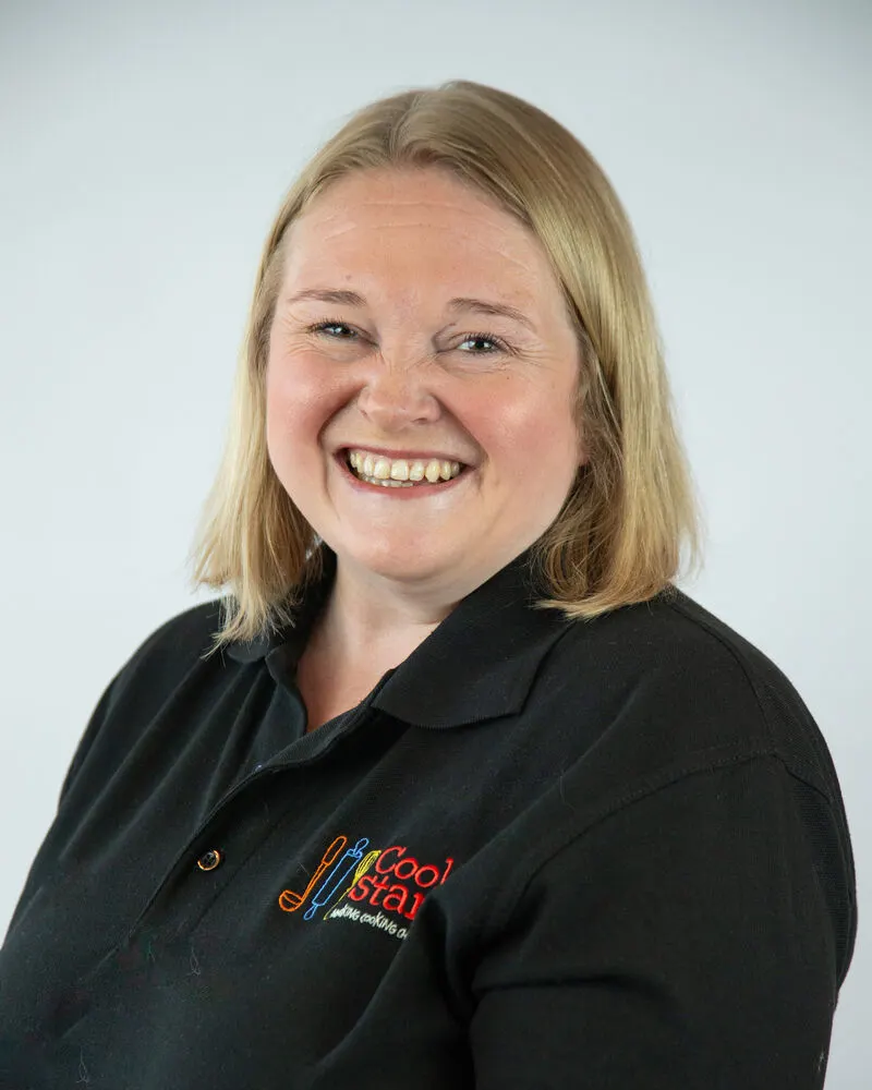 Woman with blonde hair wearing black polo shirt with venue logo, smiling at camera against white background.