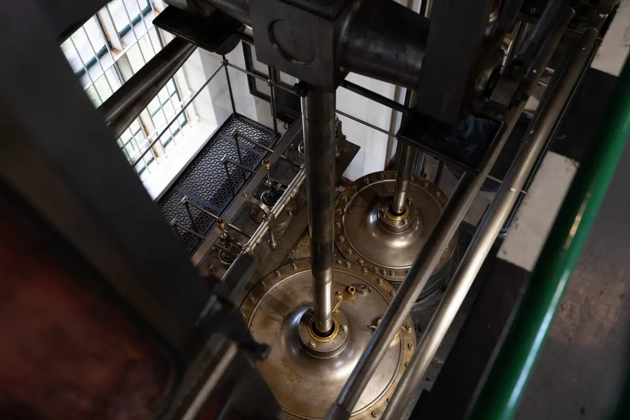 Overhead view of two large industrial steam engine cylinders with brass fittings and metal framework inside a museum building.