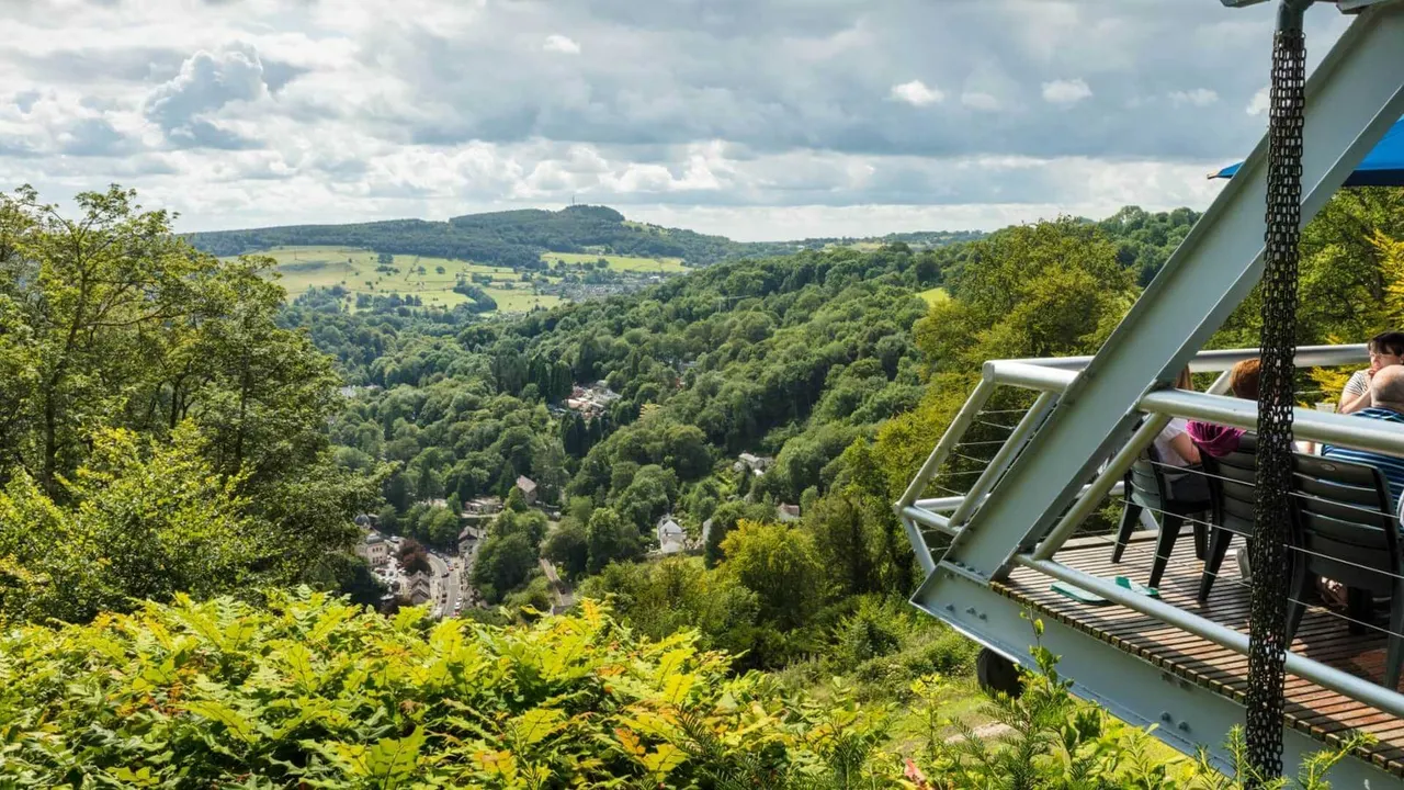 Visitors on a viewing platform overlook a vast wooded valley with distant hills and rolling countryside under cloudy skies.