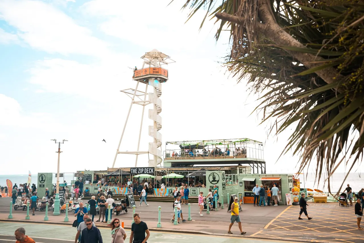 This is a PHOTOGRAPH.

Busy seaside promenade with visitors around a tall zip wire tower, green food venue building, and palm tree fronds overhead.