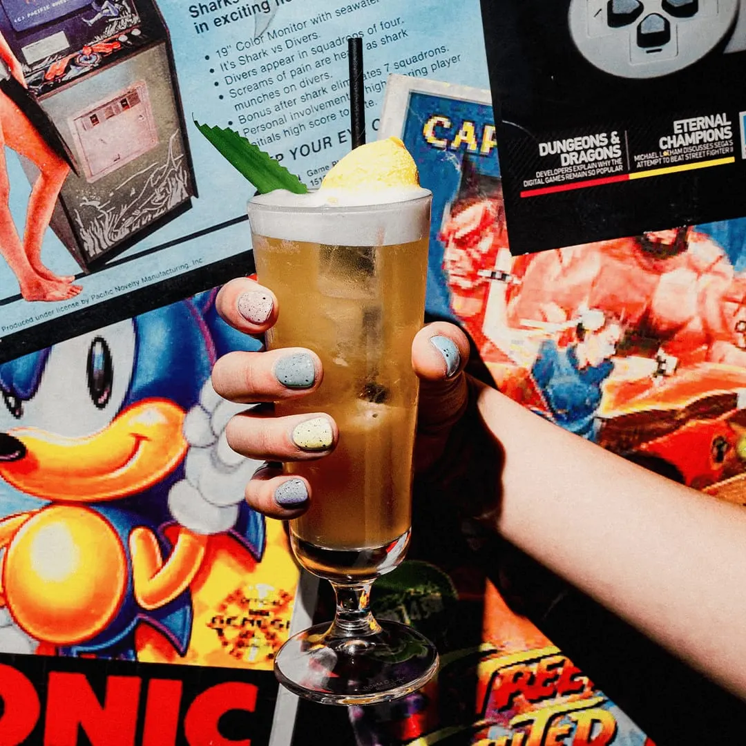 Hand holding a glass of amber beer with foam top and lemon garnish, surrounded by vintage arcade game graphics and controllers.