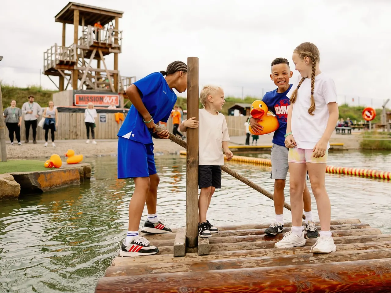 An instructor in blue helps children collect yellow ducks from water at an outdoor activity centre, with a wooden tower and spectators in the background.