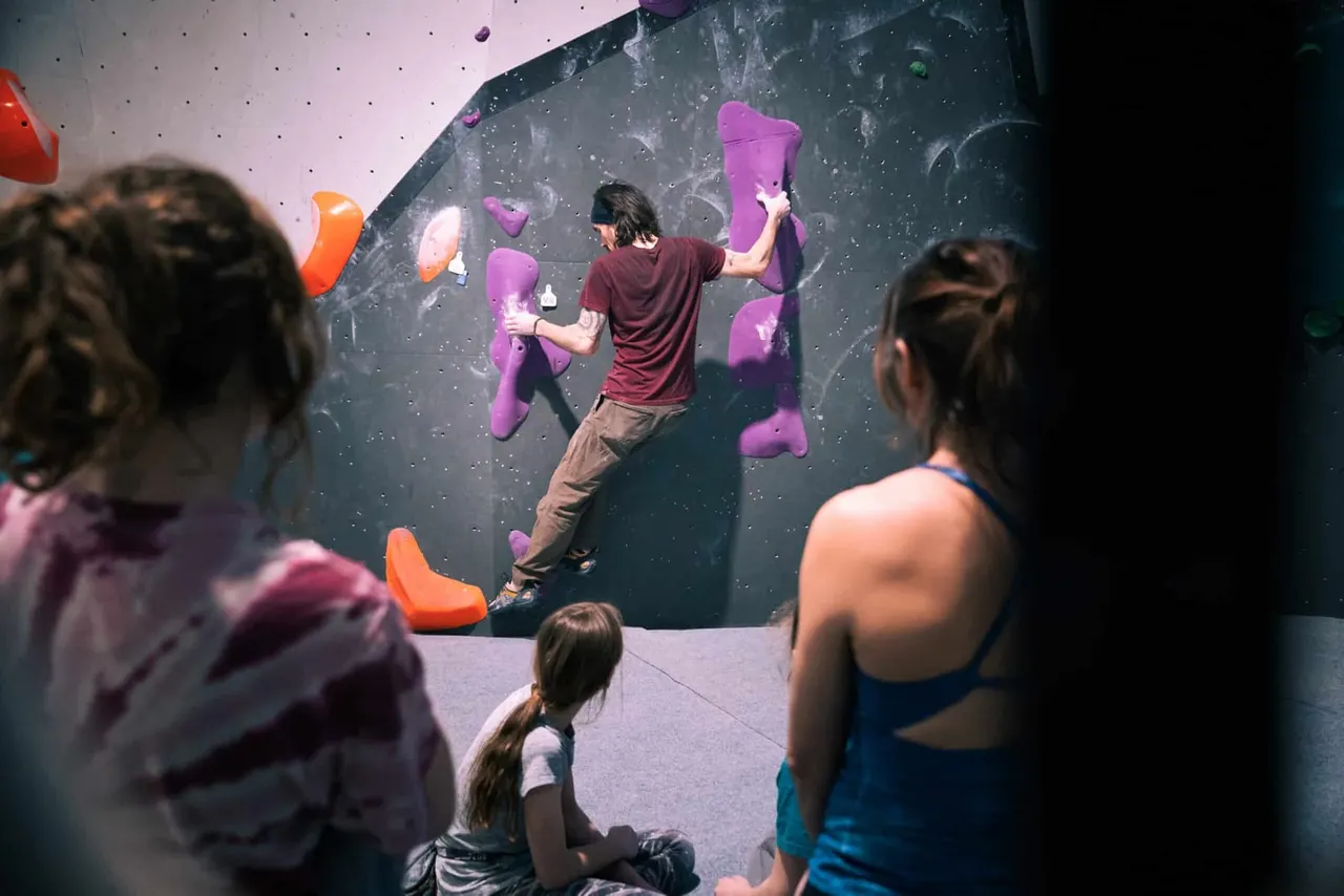 A young person indoor rock climbing on a wall with coloured holds, watched by other climbers below.