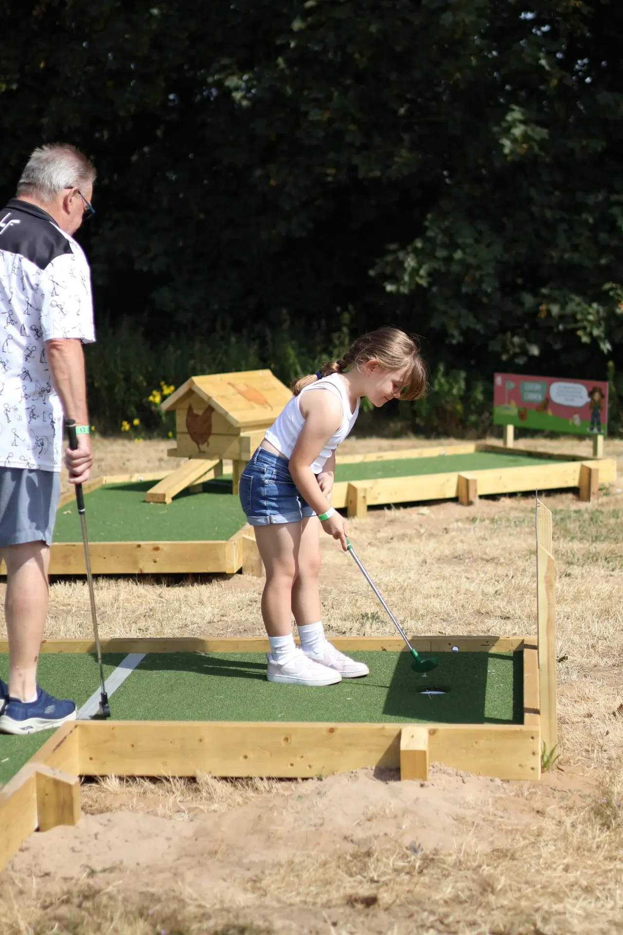 A young girl plays mini golf on an outdoor course whilst an older man watches, with wooden course features and green turf visible.