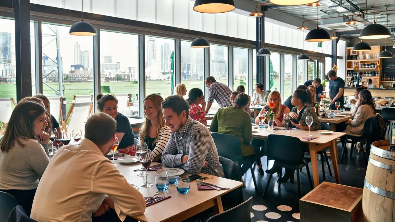 Busy restaurant with diners seated at tables enjoying meals and drinks, floor-to-ceiling windows overlooking a city skyline and green space.