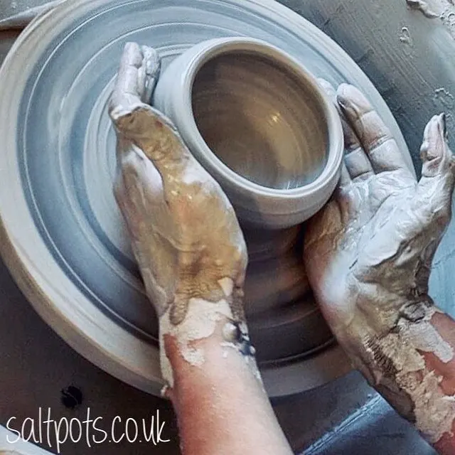 Hands covered in clay working at a pottery wheel with a ceramic vessel.