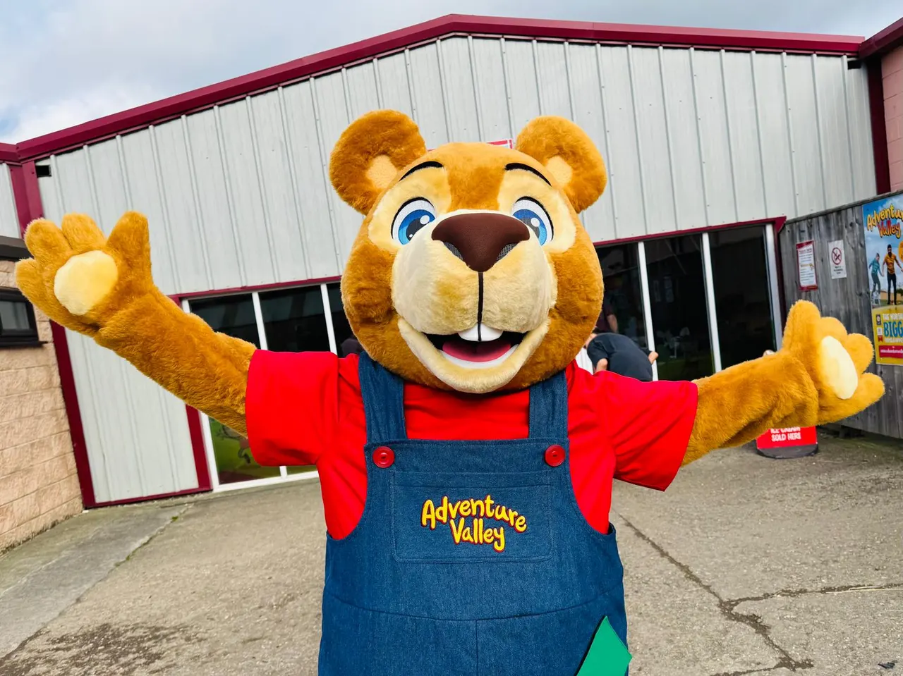 A large bear mascot in red shirt and denim overalls waves outside an adventure park building.