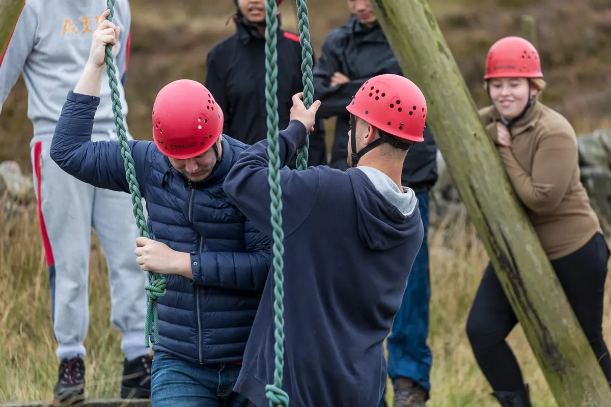 Children wearing red safety helmets climbing ropes suspended from a wooden beam outdoors.