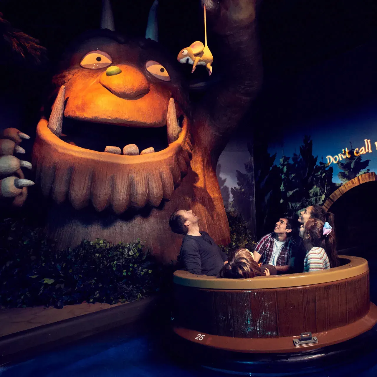 Visitors in a ride vehicle looking up at a large glowing orange monster puppet with a small creature dangling above it in a dark themed attraction.