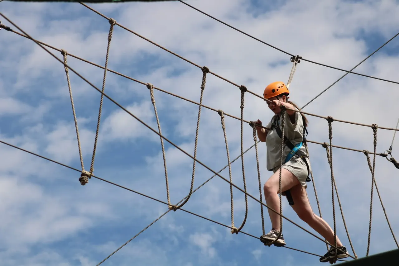 Person in orange helmet traversing a rope course obstacle against blue sky.