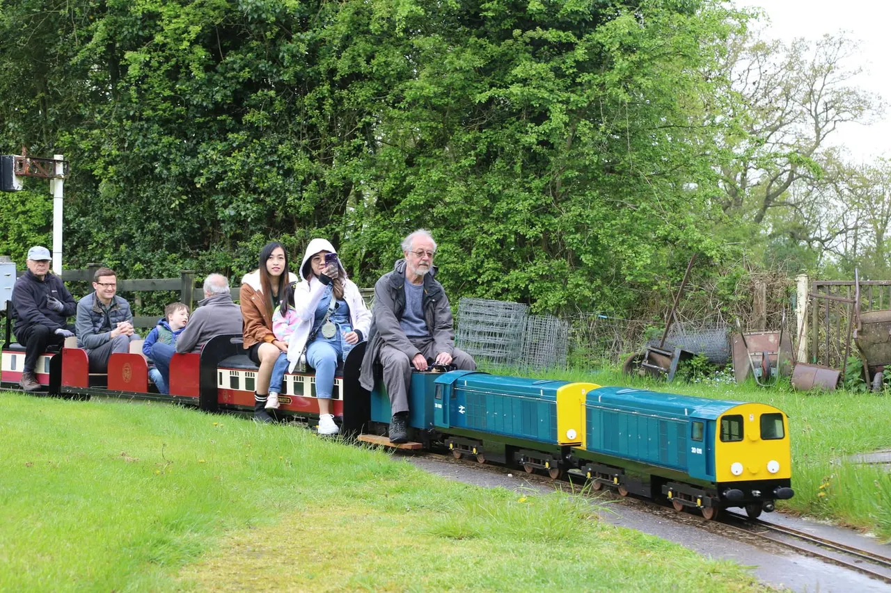Families and children riding on a miniature railway train through a green garden setting.