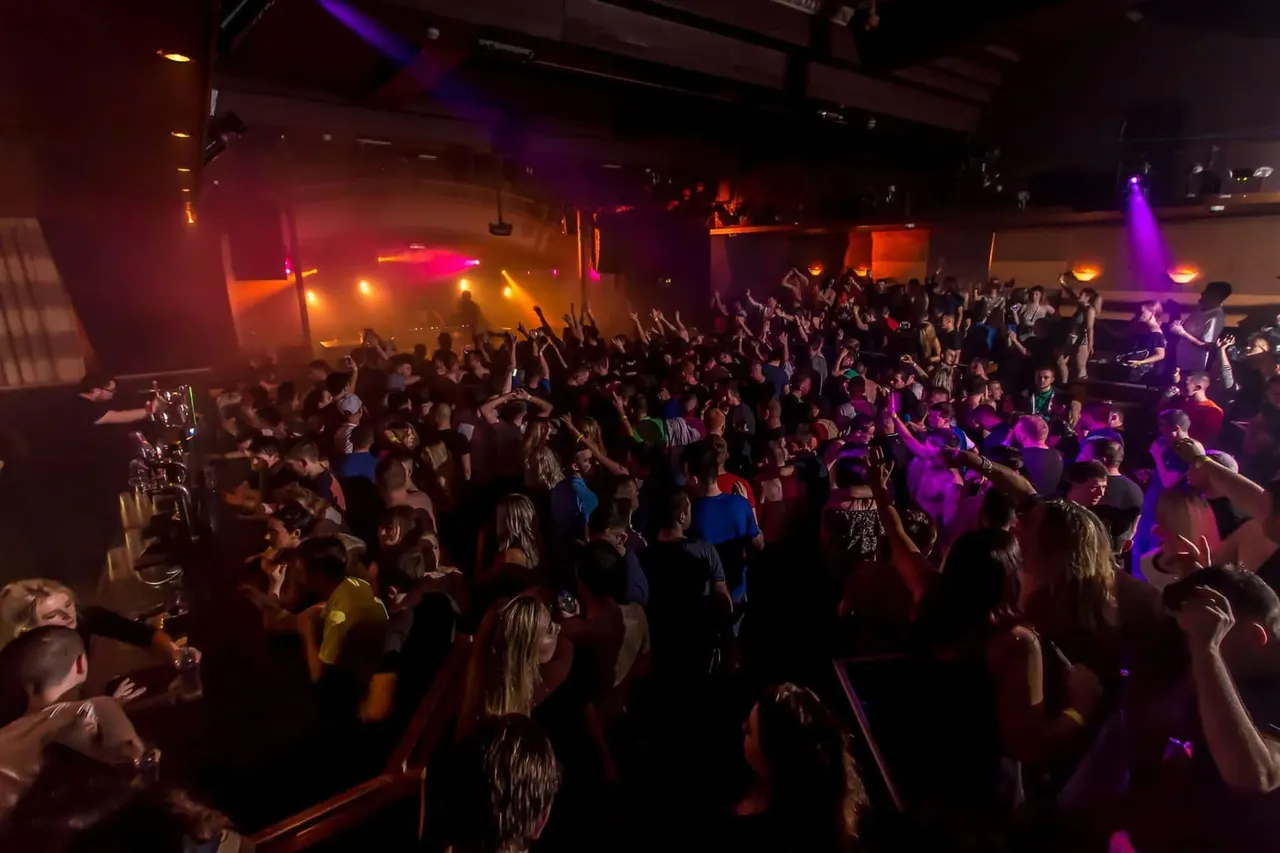Large crowd of people dancing and celebrating in a nightclub with colourful stage lighting.