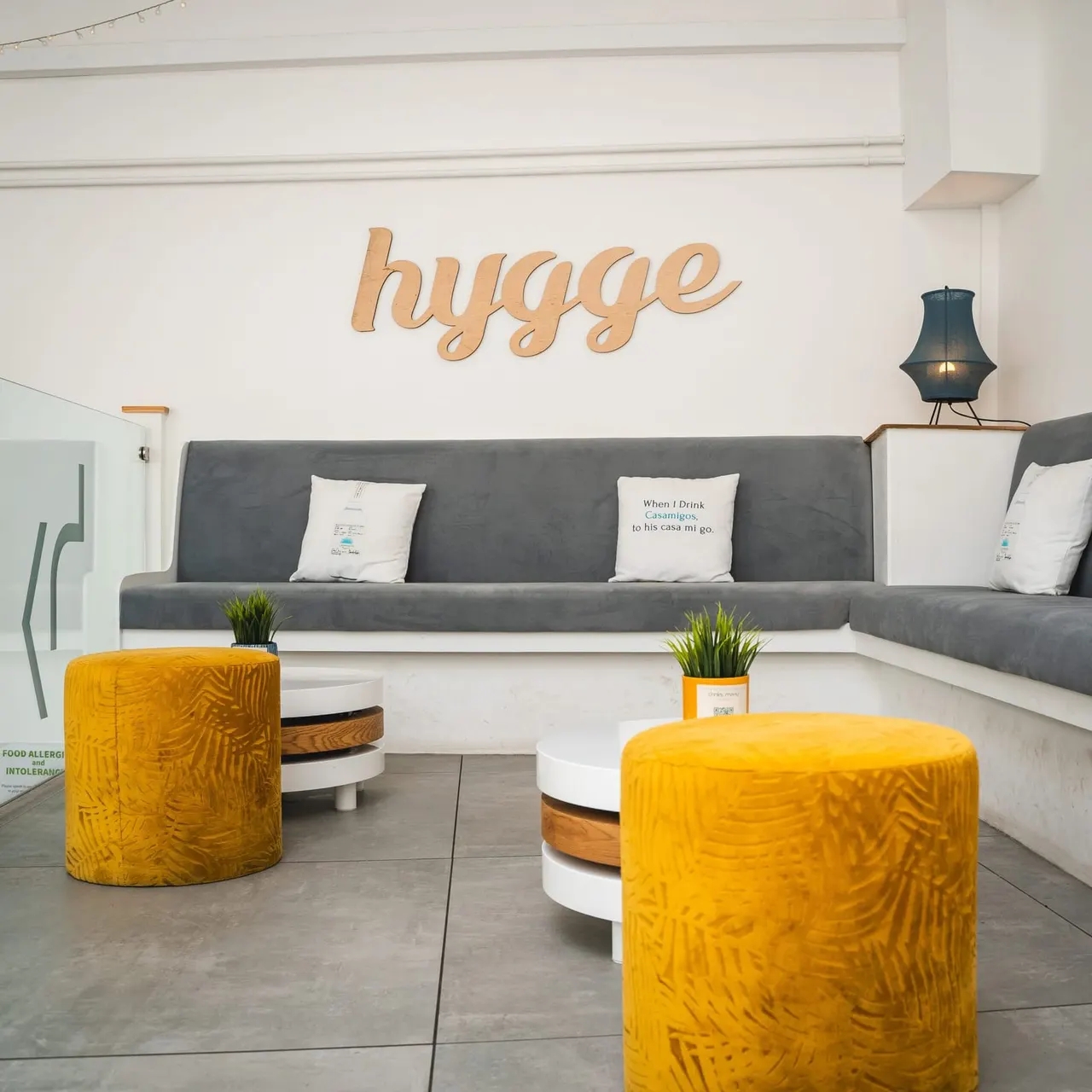 Modern lounge interior with grey upholstered seating, yellow patterned poufs, potted plants and wooden "hygge" wall sign.