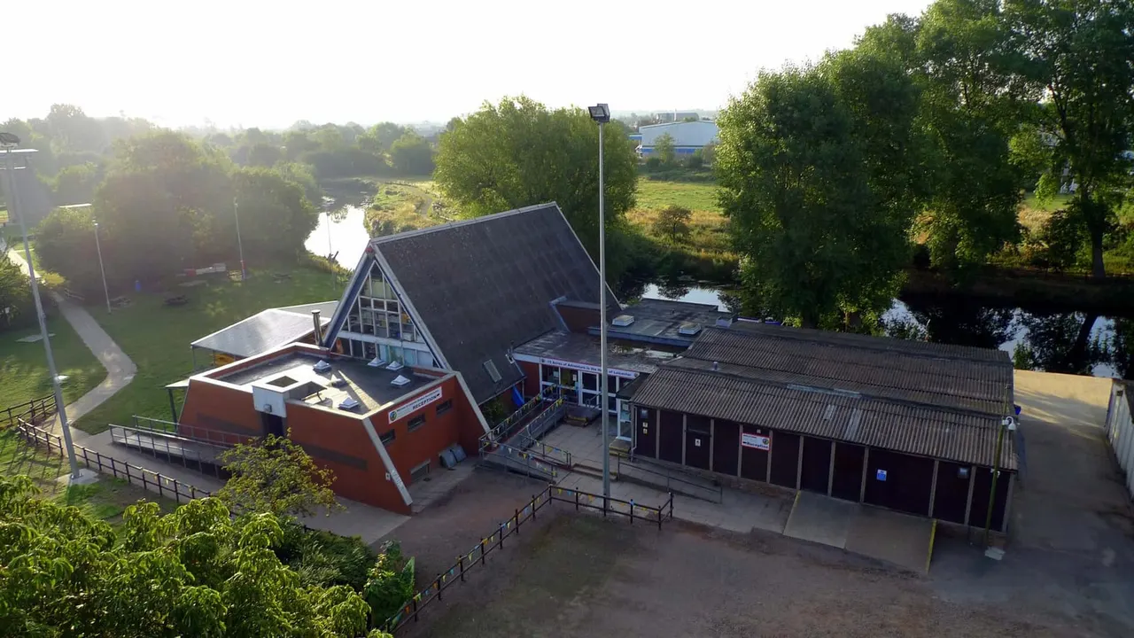 Aerial view of a leisure venue with distinctive red building and dark-roofed structures beside a river, surrounded by trees and green fields.