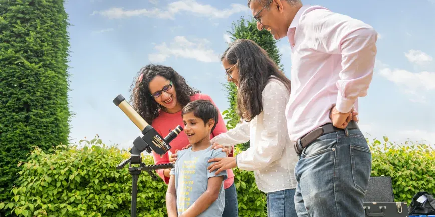 Family of four using a telescope in a garden surrounded by tall hedges on a sunny day.