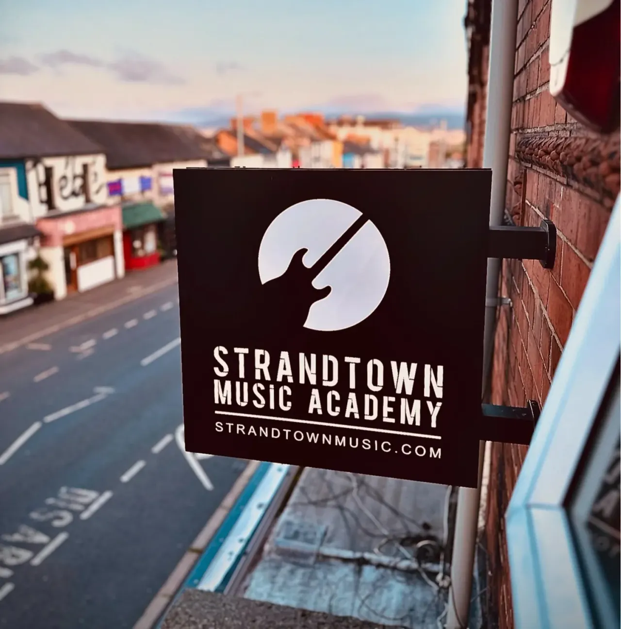 Strandtown Music Academy signage mounted on a brick building with a street and hills visible in the background.
