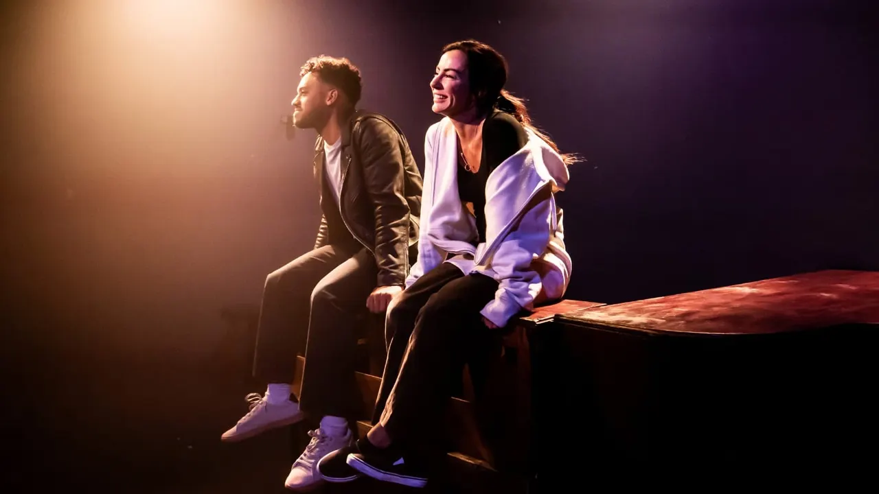 Two performers sit on stage under warm and purple lighting during a theatrical production.