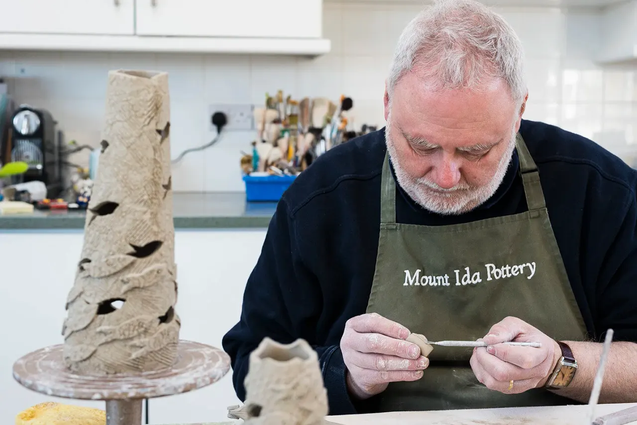 Potter working on clay sculpture in studio workshop, with carved ceramic pieces on table beside him.