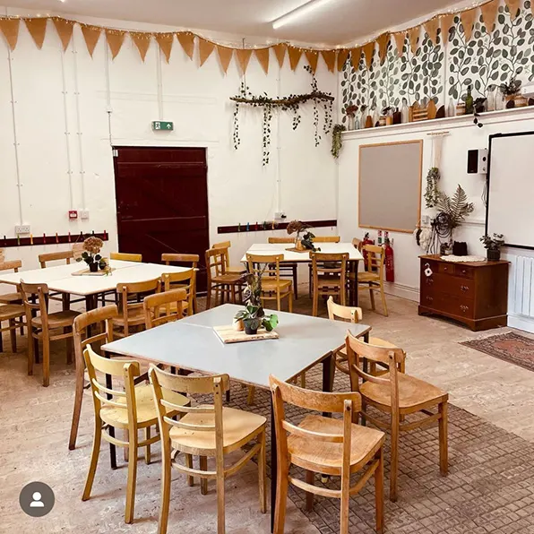 Interior of a decorated hall with multiple wooden tables and chairs arranged for gatherings, festive bunting and garland decorations overhead.